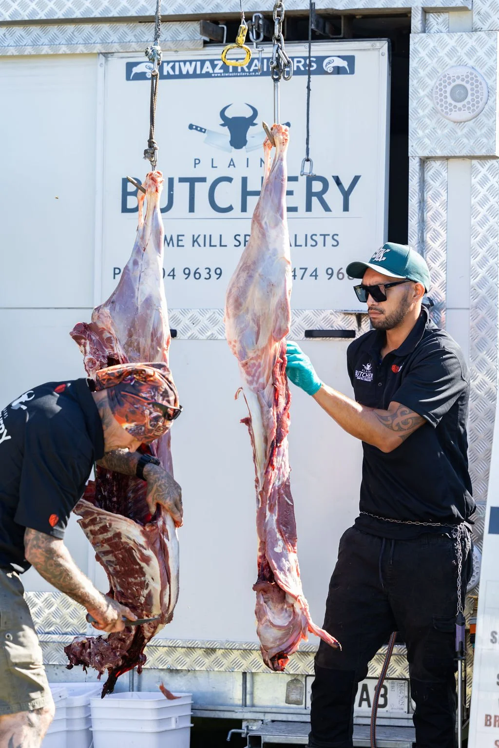 Two butchers handling large carcasses of meat hanging from chains outside a butchery shop. One butcher is wearing sunglasses and gloves, examining a carcass, while the other is cutting meat with a knife.