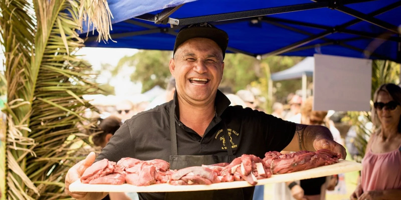 A man smiling and holding a large tray of raw meat at an outdoor market or event, with people in the background.