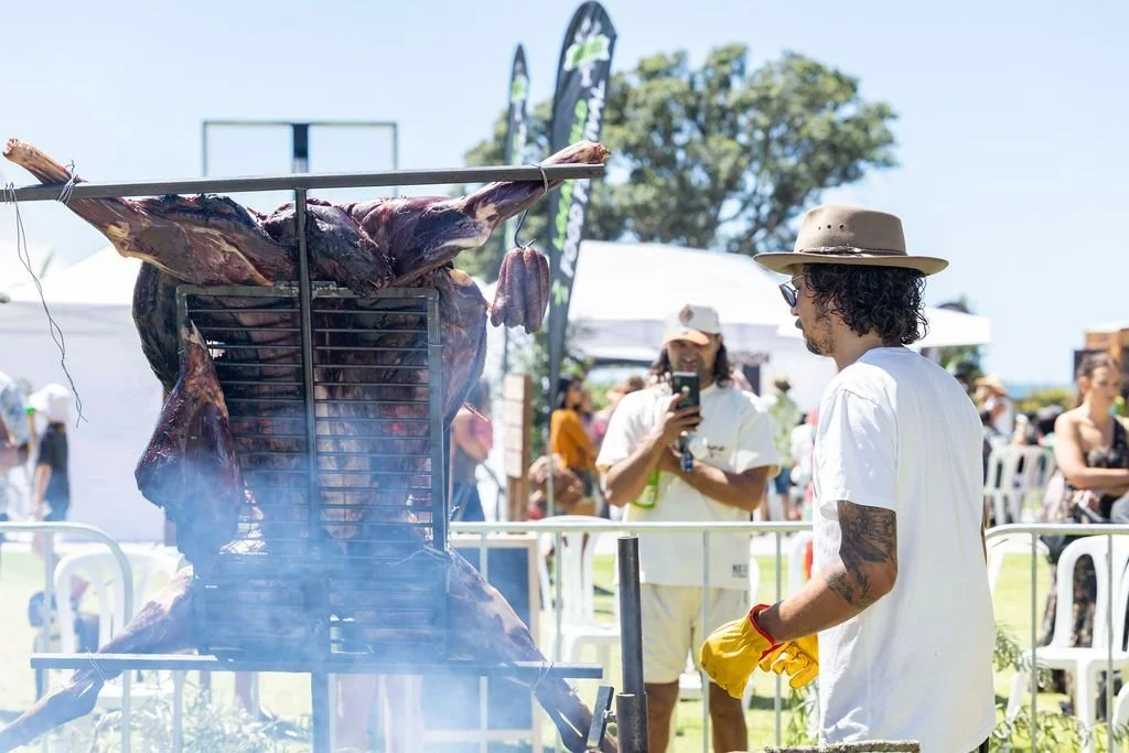 A person grilling a whole animal on a spit at an outdoor event, with other people and tents in the background.