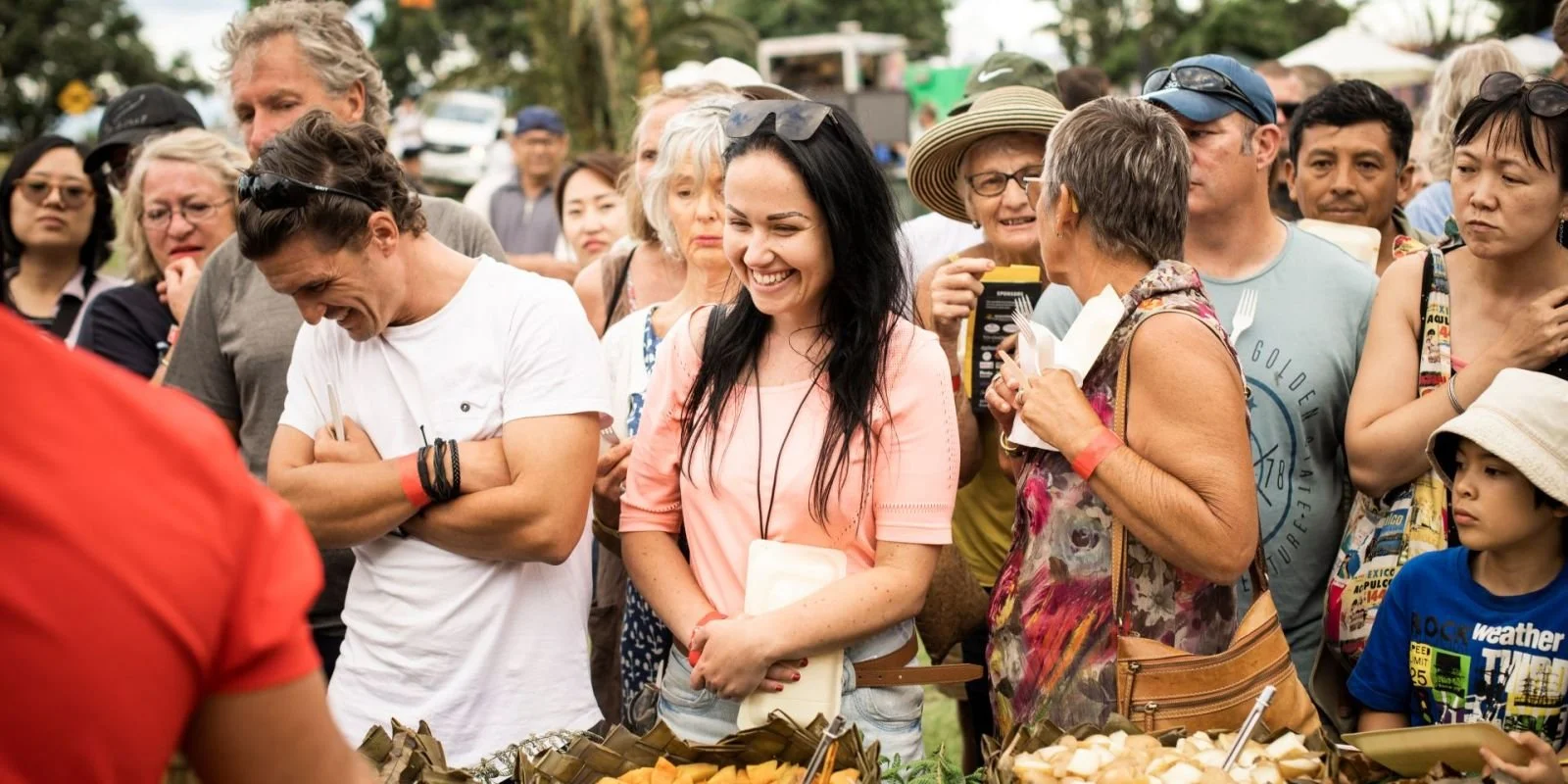 Crowd of diverse people gathered outdoors, smiling and engaging, with food in the foreground.