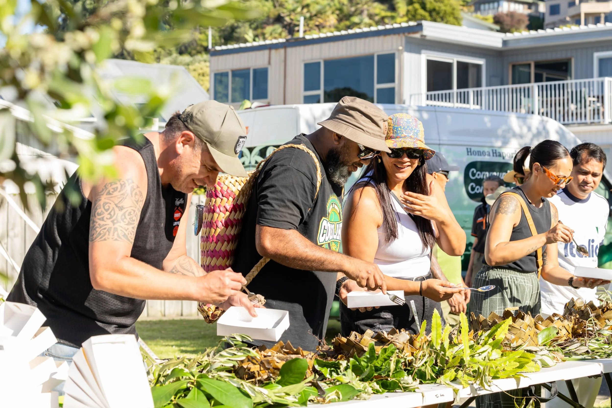 A group of diverse people are gathered around a table outdoors, eating food served in takeout boxes. They are smiling and appear to be enjoying a communal meal at a sunny event.