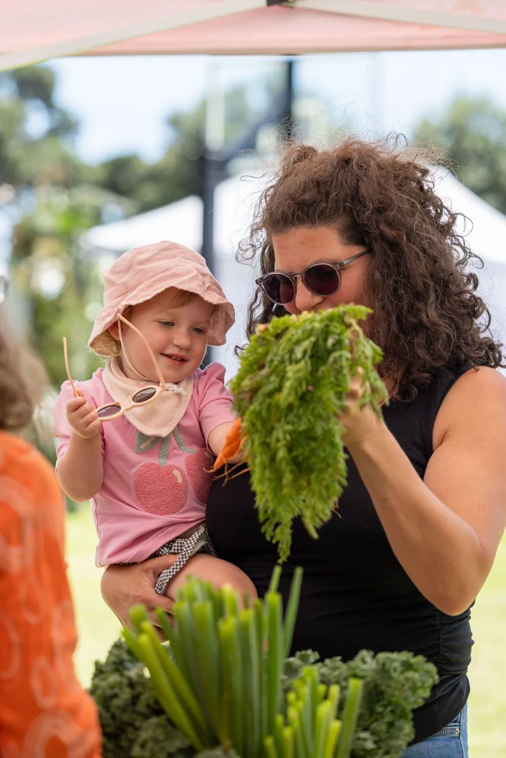 A woman holding a young girl at a market or farm stand, examining fresh vegetables, with other children and tables with produce in the background.