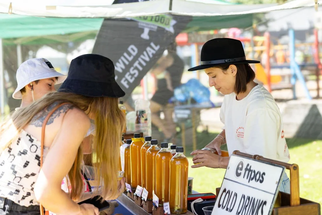 A woman working at a beverage stand selling cold drinks at an outdoor festival with a 'Wild Festival' sign in the background, and three young women buying drinks.