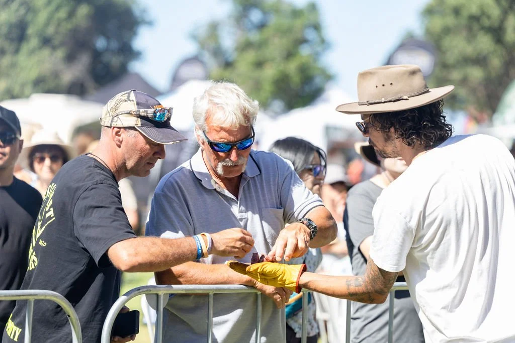 Three men at an outdoor event, one wearing a black T-shirt and cap, another with white hair and sunglasses, and the third with curly hair, a hat, and a white T-shirt, exchanging an fire cooked meat.