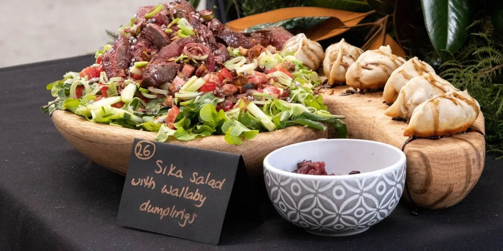 A wooden platter with Sika salad with wallaby dumplings and a small bowl of wallaby meat. The salad contains chopped vegetables and wallaby meat pieces, with dumplings on the side. A black sign indicates "Sika salad with wallaby dumplings."