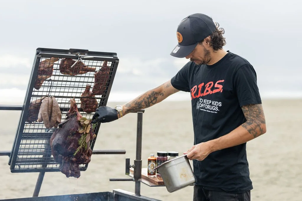A man with tattoos wearing a black T-shirt and a black baseball cap prepares meat on a grill at the beach.