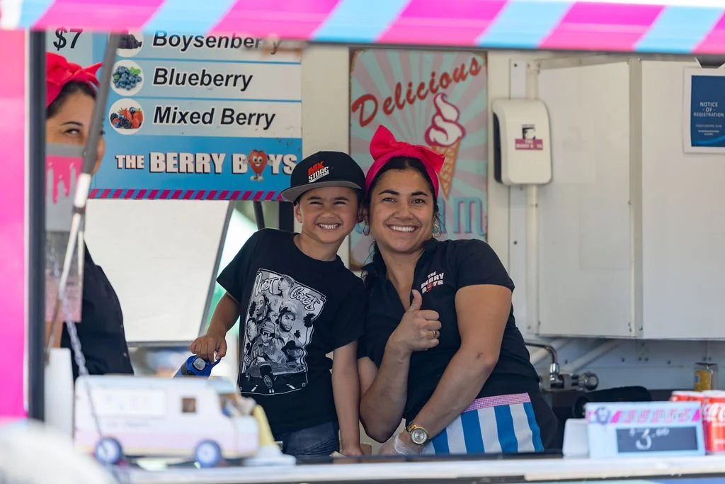 A smiling woman with a pink headband giving a thumbs-up and a young boy with a black T-shirt and a cap, both standing inside a colorful ice cream stand. The stand has signs advertising various berries and a pink and blue striped banner. The woman is wearing a black shirt with a logo, and the boy is holding a blue item, possibly a toy or food item.