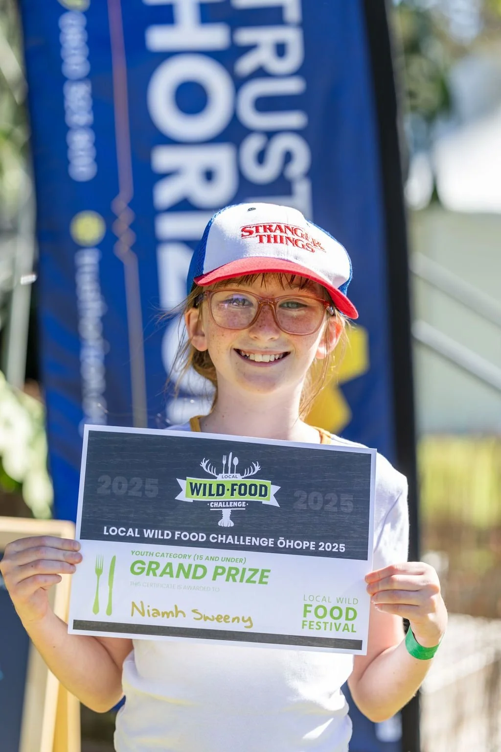A young girl with glasses and freckles smiling, holding a certificate for the Grand Prize at the 2025 local wild food challenge, standing outdoors during the food festival.
