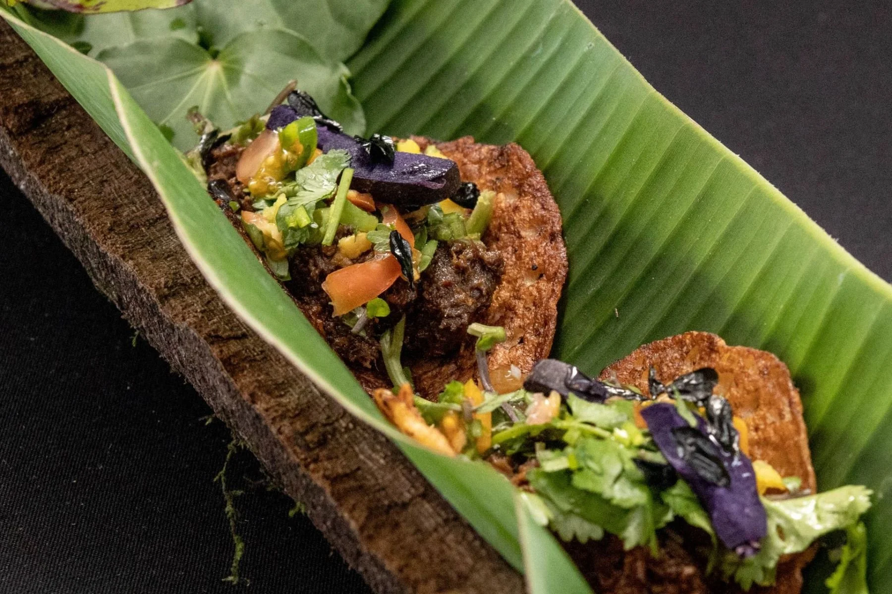 Fried meat topped with chopped vegetables and herbs, served on a green banana leaf lined on a rustic wooden serving plank.