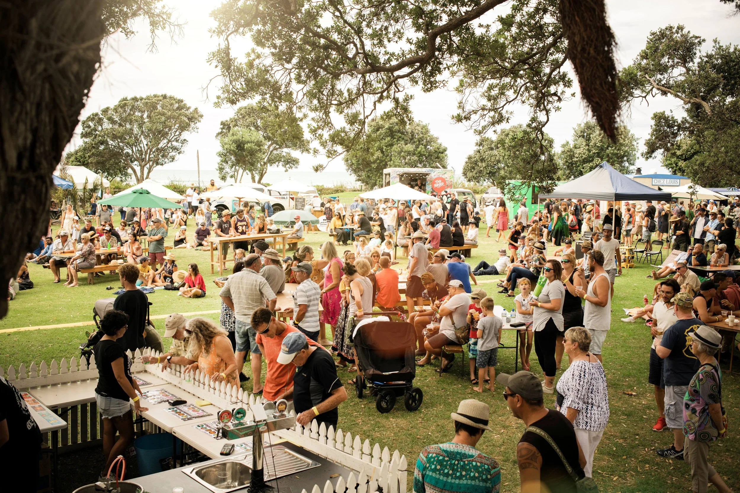 A large outdoor festival with many people gathered on a grassy lawn, some sitting on benches and others standing. There are tents and booths in the background, with some trees and a stage visible.
