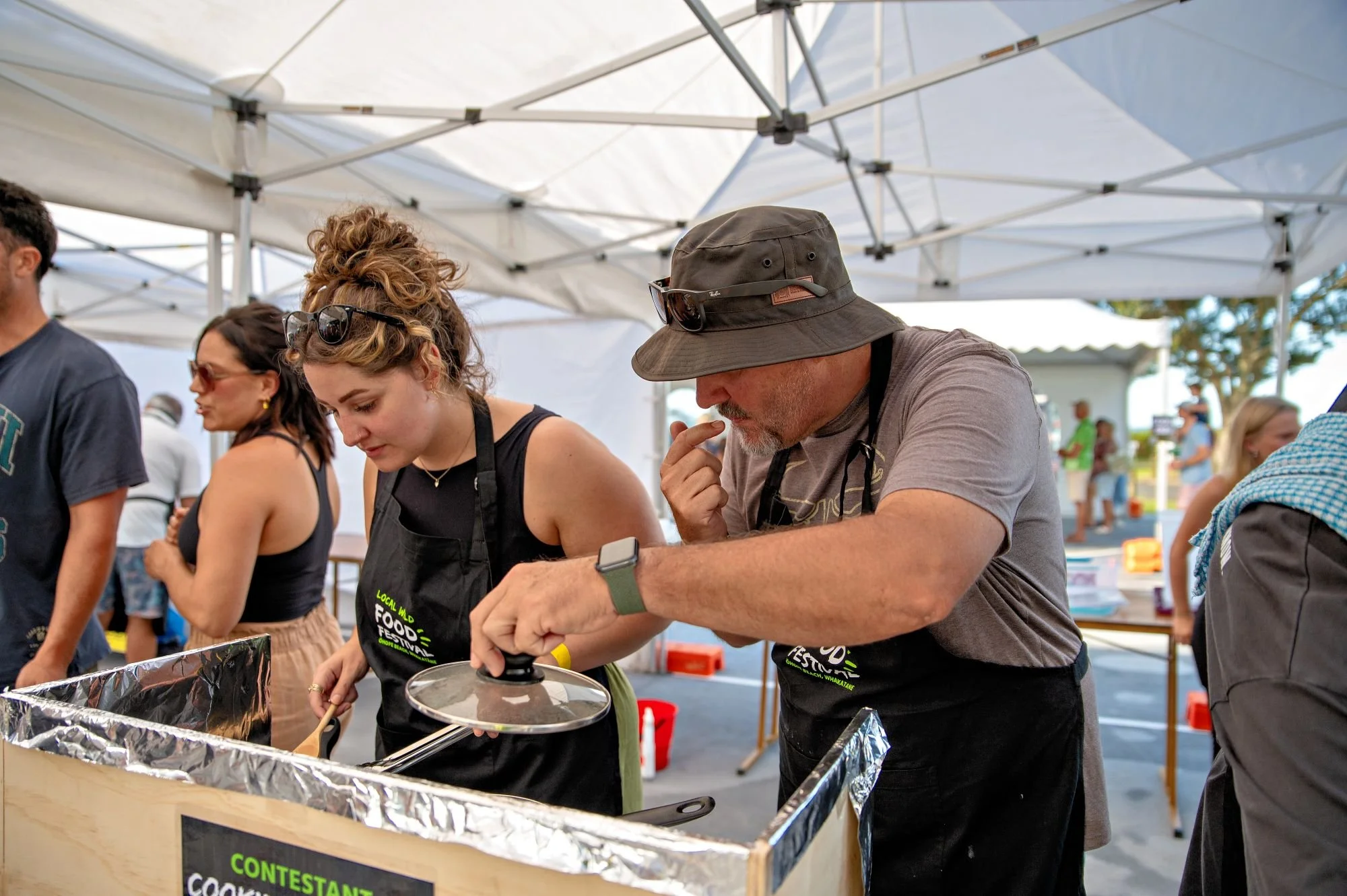 Two people cooking at a food festival stand under a white canopy, with other festival attendees in the background.