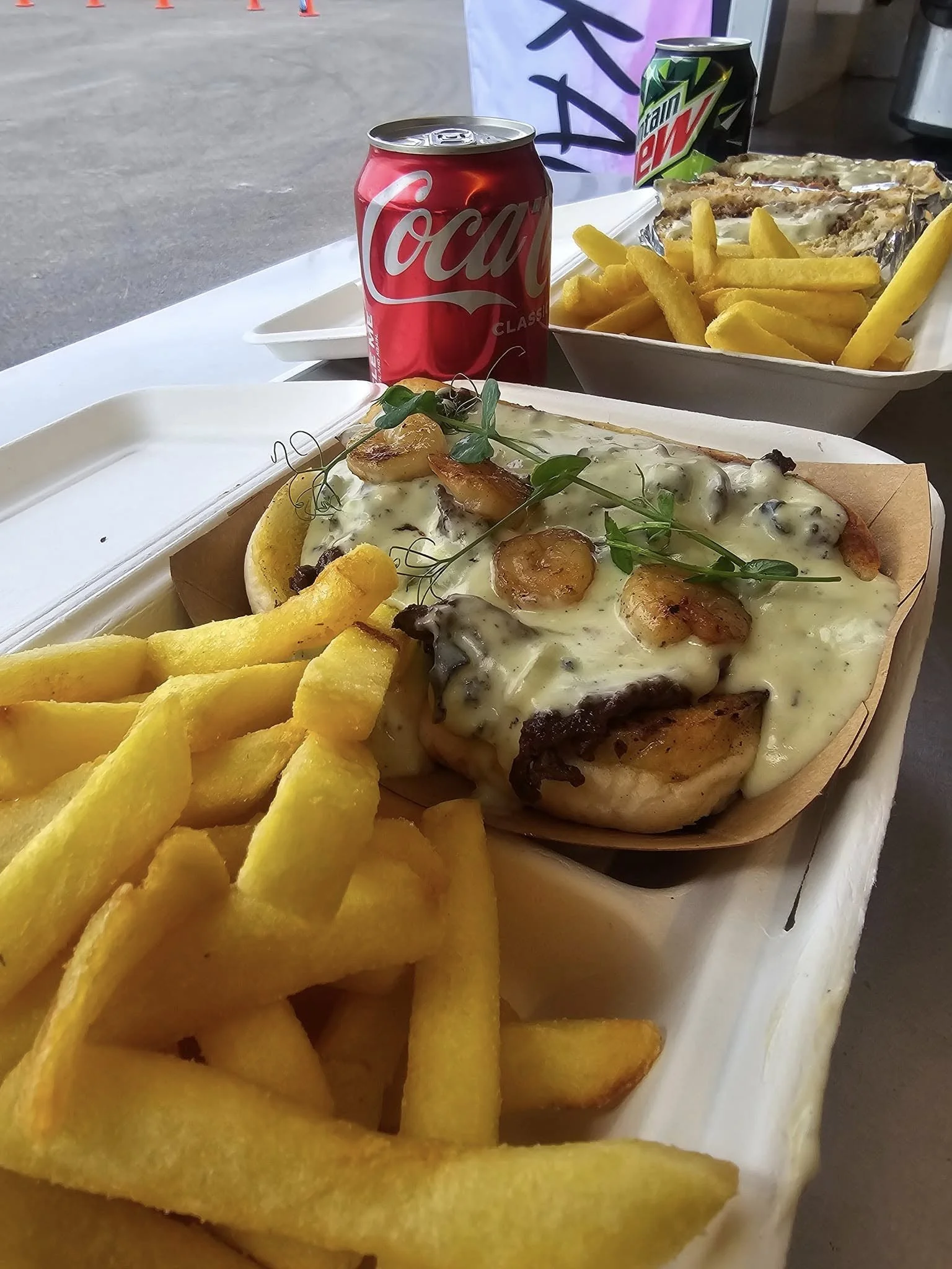 Creamed paua and grilled shrimp, and microgreens, served with French fries and two cans of soda, Coca-Cola, on a white tray.