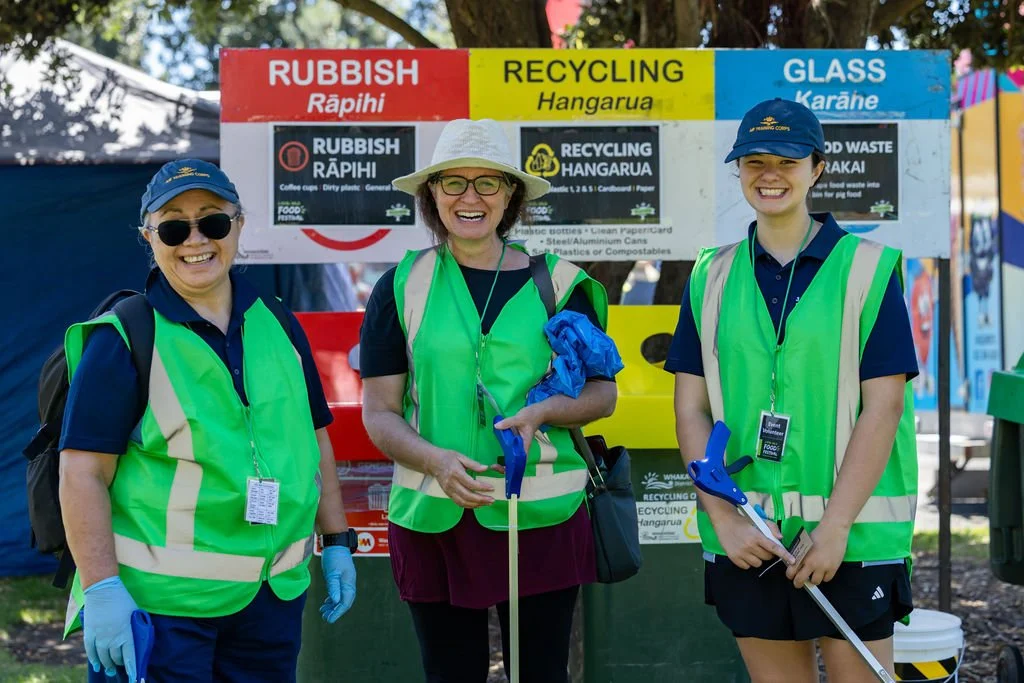 Three women in safety vests and hats smiling at a recycling station with signs for rubbish, recycling, and glass.