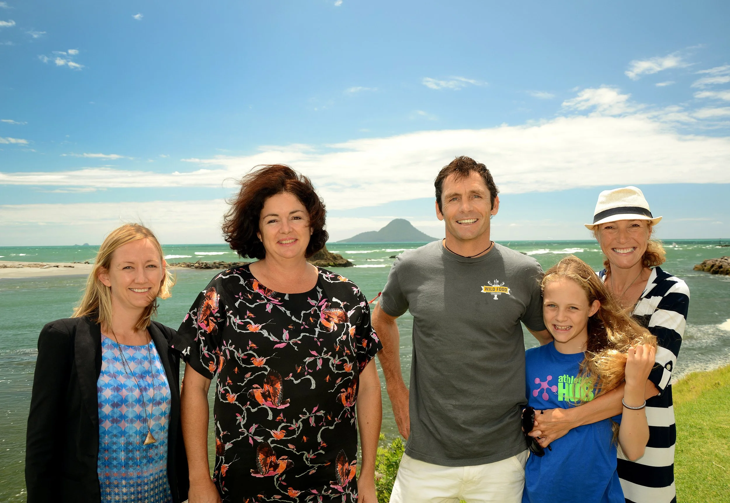 Group of six people standing outdoors near the ocean with a small island in the background, smiling at the camera.