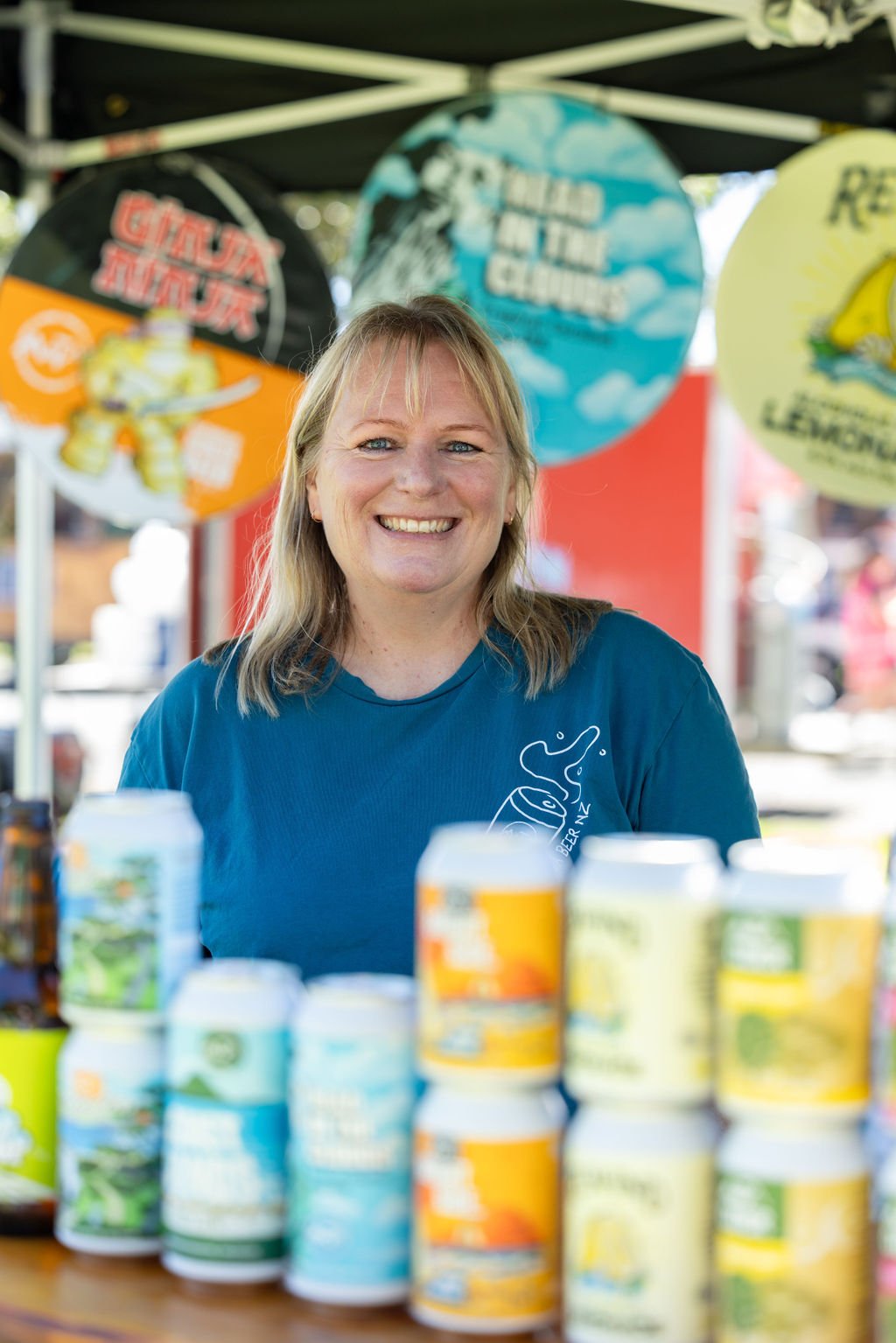 Smiling woman at an outdoor booth with beer cans and colorful signs in the background.