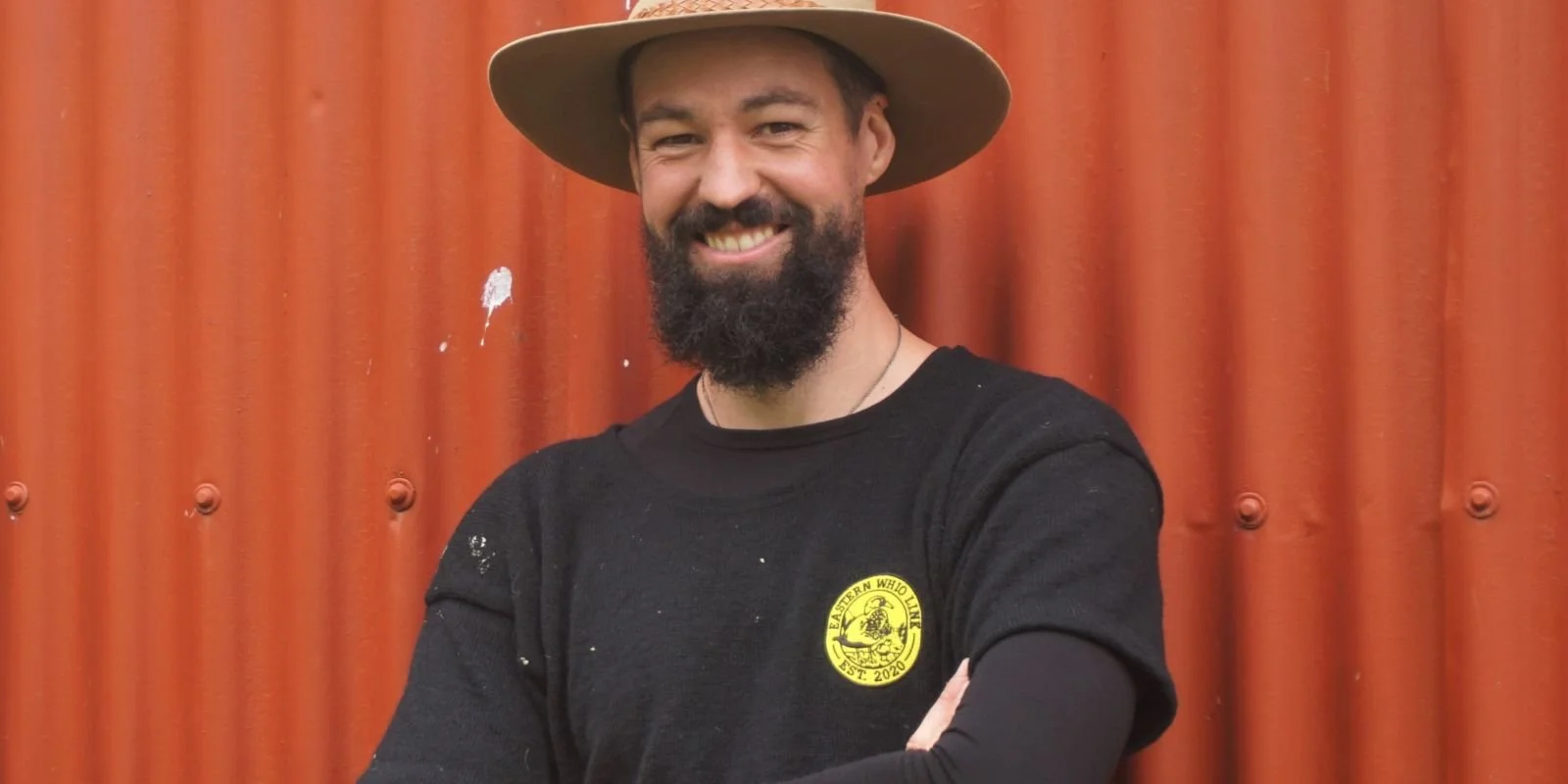 Photo of Sam the Trapman: A man with a beard and mustache wearing a wide-brimmed hat and a black T-shirt with a yellow circular logo, standing in front of a red corrugated metal wall. He is smiling and has his arms crossed.