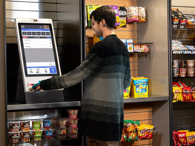 A person using a self-checkout kiosk in a grocery store, surrounded by shelves of snacks and convenience foods.