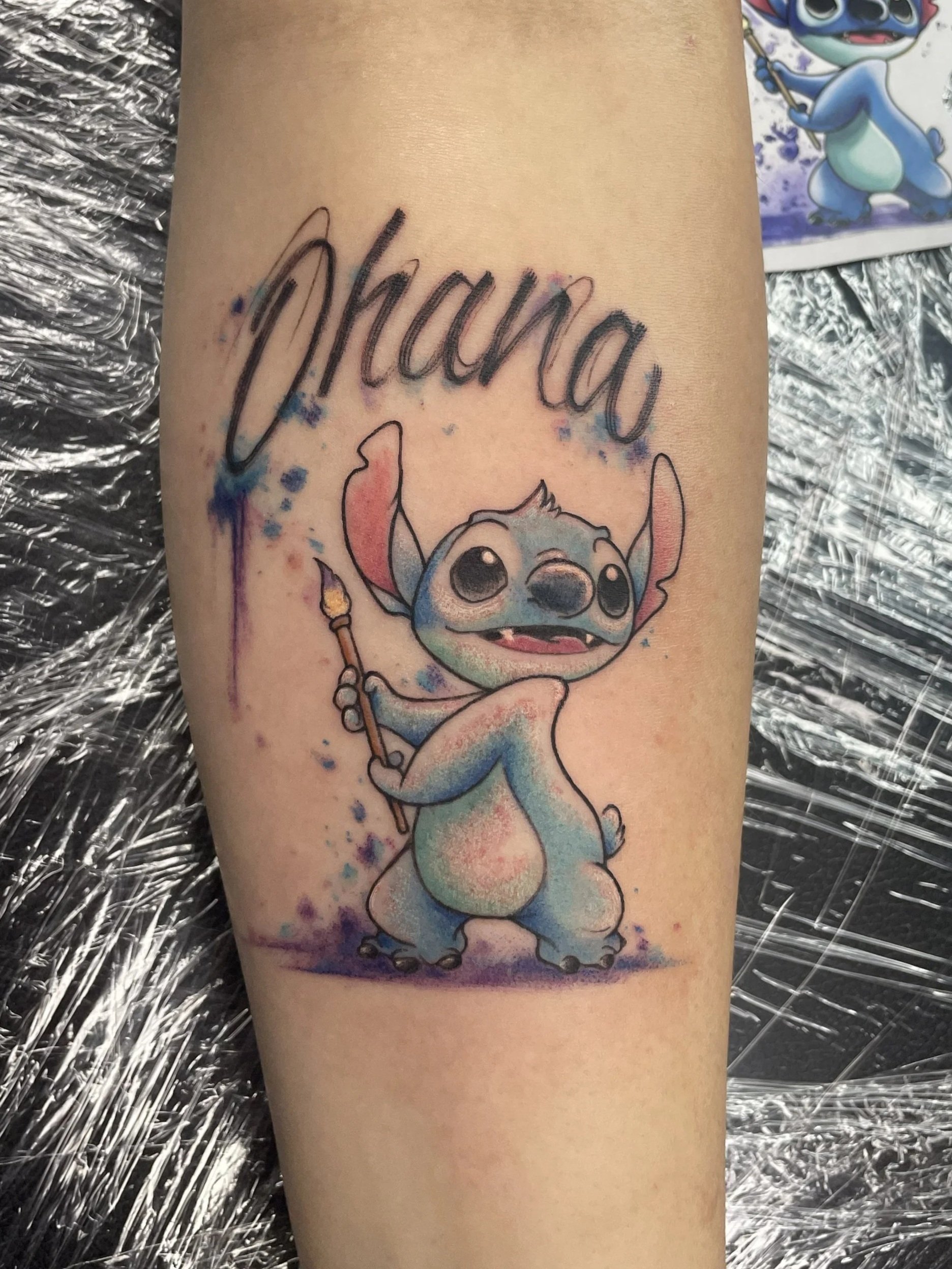Tattoo of a cartoon character, Stitch from Disney's Lilo & Stitch, holding a paintbrush, with "OHANA"  written above in cursive, tattooed by Zing Phelps, in Cedar Rapids, Iowa