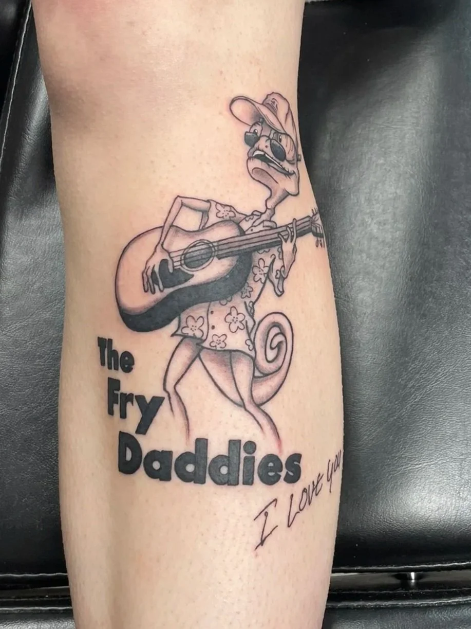 Tattoo of a lizard wearing a hat and sunglasses, playing a guitar, with the text "The Fry Daddies" and a handwritten "I Love You", tattooed by Zing Phelps, in Cedar Rapids, Iowa