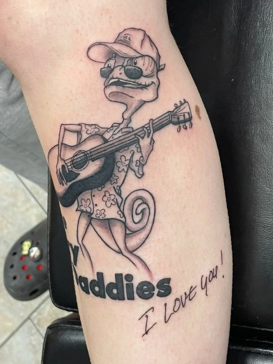 Tattoo of a cartoon lizard wearing a cap, playing an acoustic guitar, tattooed by Zing Phelps, in Cedar Rapids, Iowa