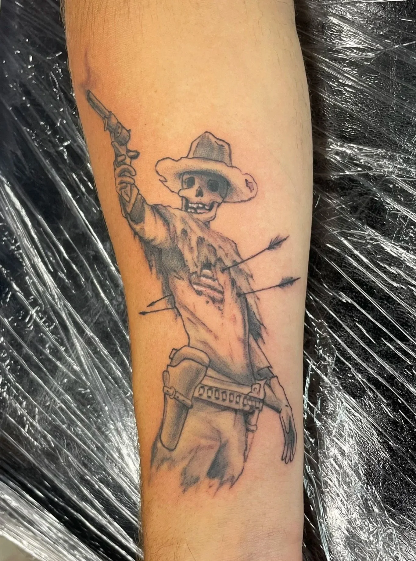 Tattoo of a skeleton cowboy, holding a gun in one hand, wearing a wide-brimmed hat and a tattered poncho, tattooed by Zing Phelps, in Cedar Rapids, Iowa