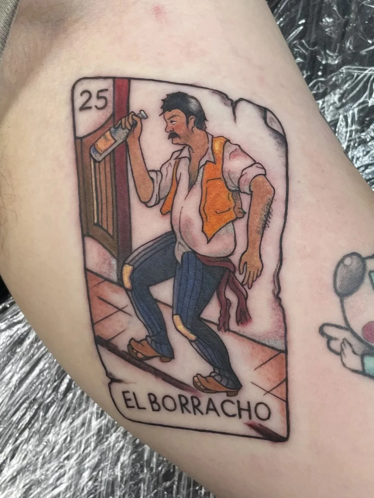 Tattoo of a cartoonish drunk man, wearing a partially unbuttoned shirt and jeans, holding a bottle, with the word 'ELBORRACHO' beneath it, tattooed by Zing Phelps, in Cedar Rapids, Iowa