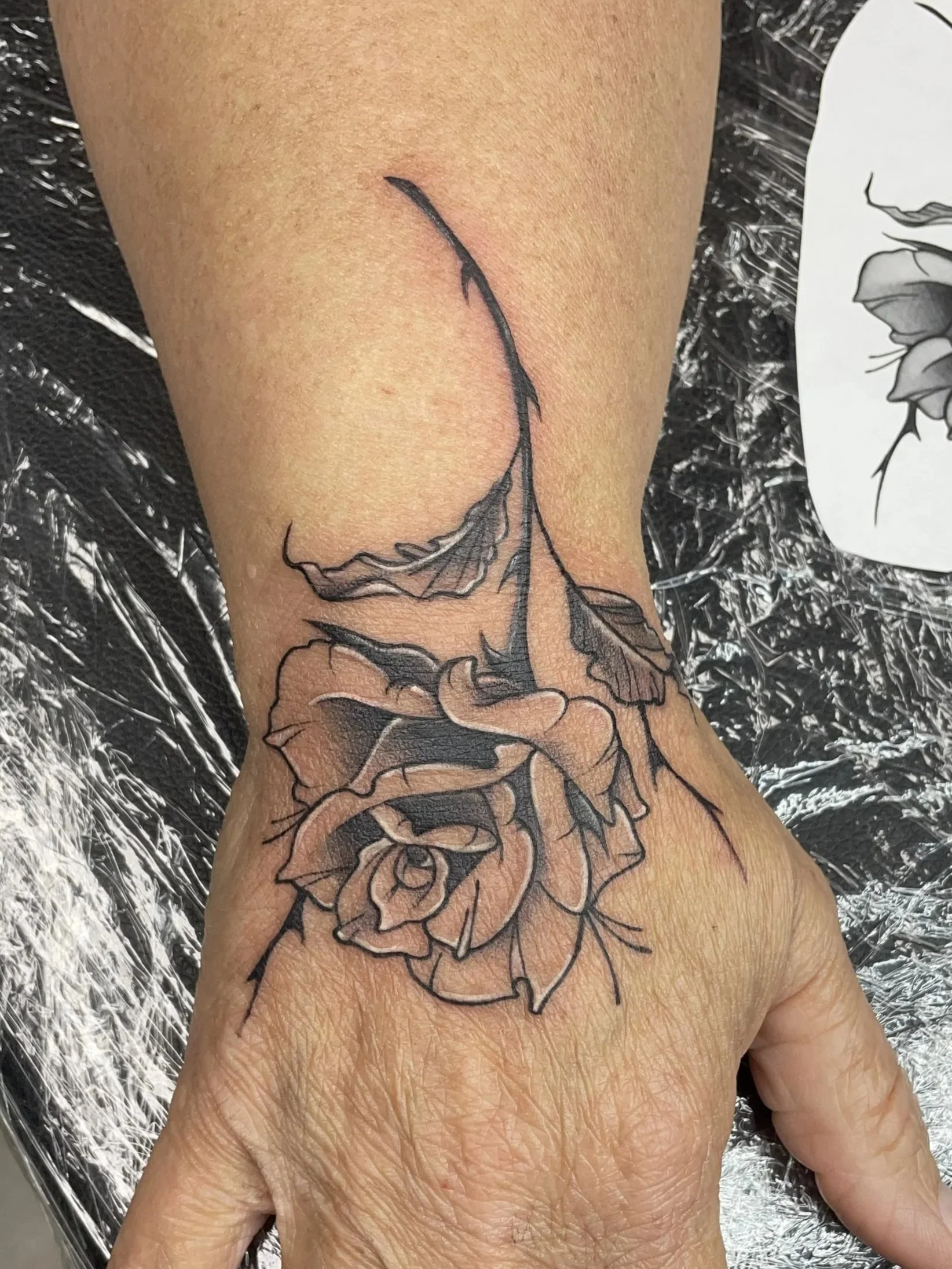 A hand with a black and gray tattoo of a rose with detailed petals and leaves, tattooed by Zing Phelps, in Cedar Rapids, Iowa