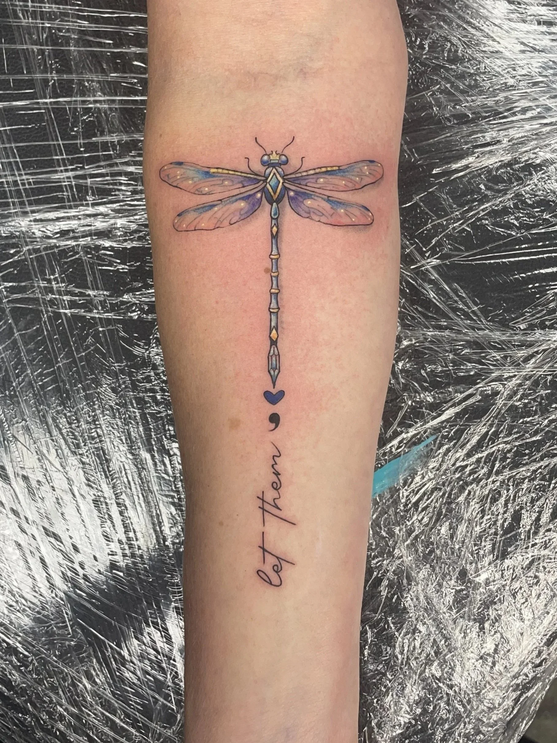 Colorful tattoo of a dragonfly with blue and purple wings on person's forearm. Below the dragonfly, the phrase "Let them" is written in cursive with a semicolon and heart symbol in blue, tattooed by Zing Phelps, in Cedar Rapids, Iowa