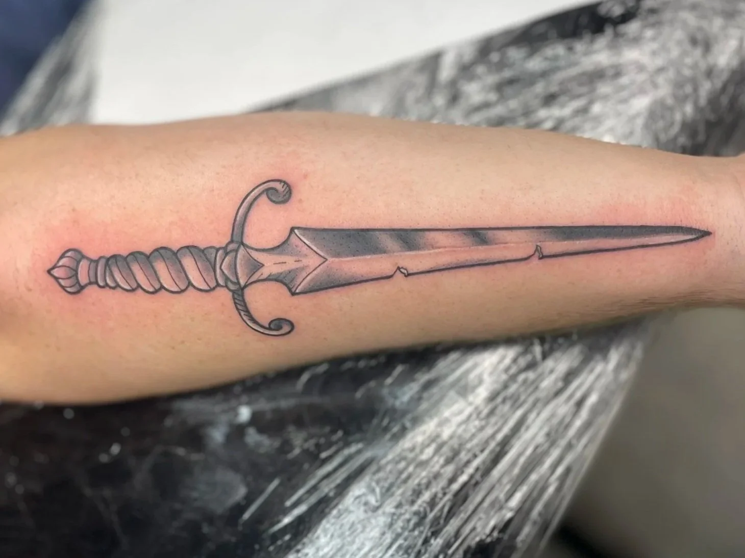 Tattoo of a detailed sword with an ornate hilt on an outer forearm, tattooed by Zing Phelps, in Cedar Rapids, Iowa
