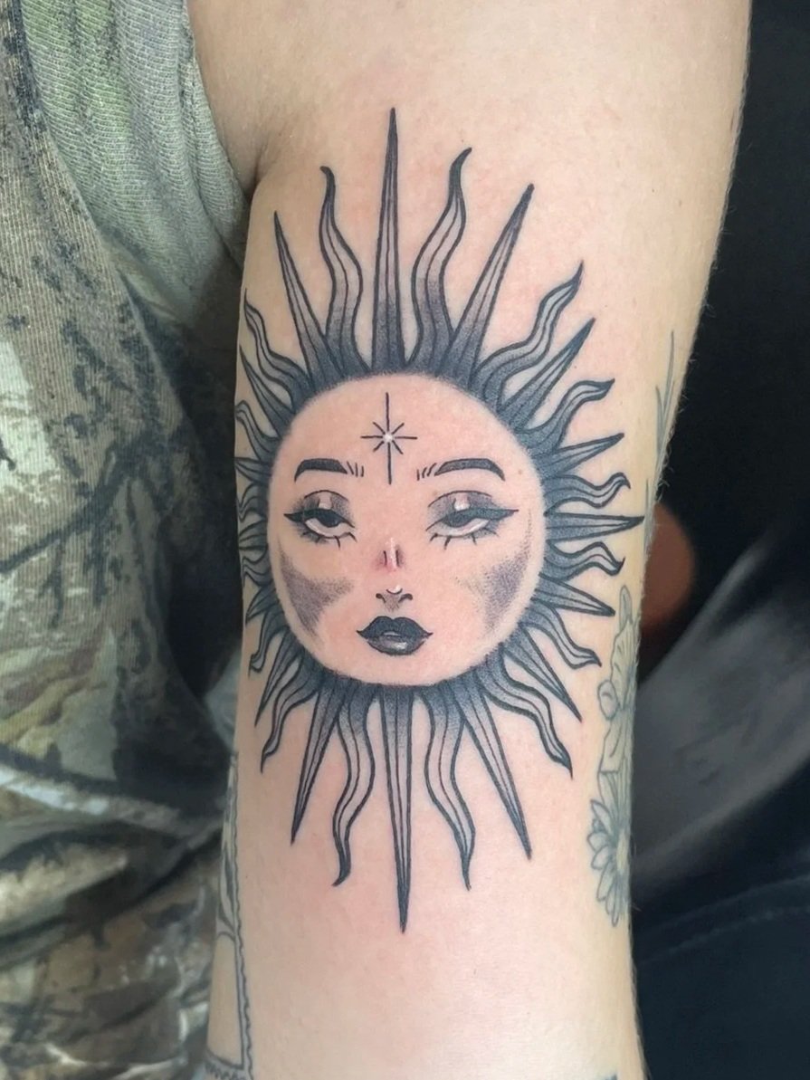 Tattoo of a stylized sun with a human face, dark makeup, and flowing sun rays, tattooed by Zing Phelps, in Cedar Rapids, Iowa