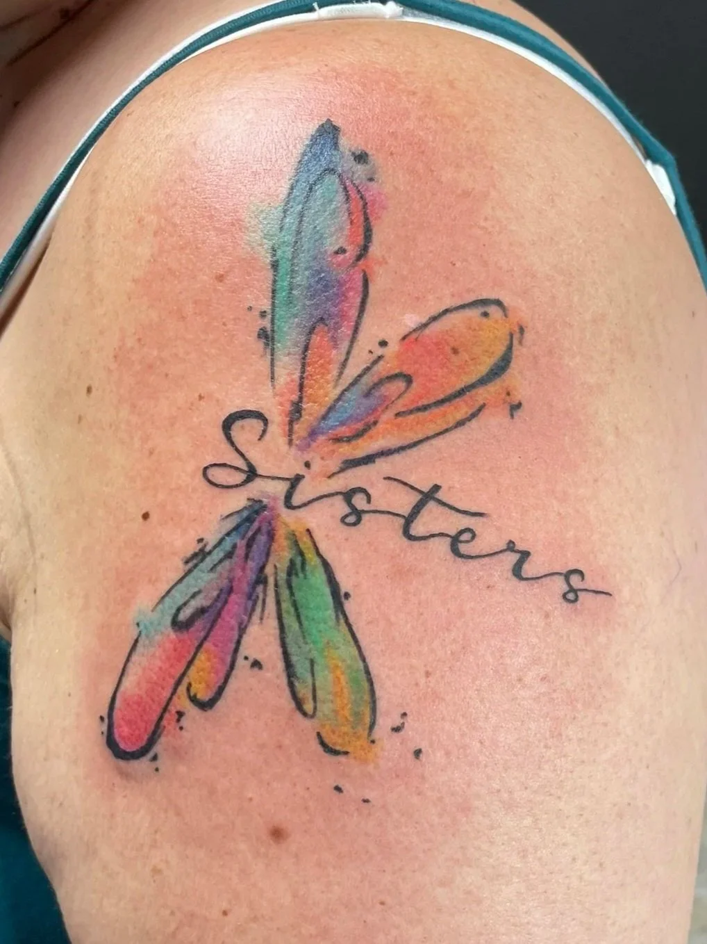 Colorful watercolor-style tattoo of a dragonfly with the word 'Sister' incorporated into the design, tattooed by Zing Phelps, in Cedar Rapids, Iowa