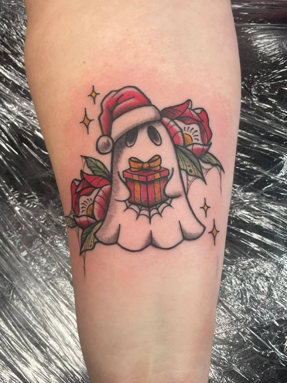 A tattoo of a ghost wearing a Santa hat, holding a gift box with a bow, surrounded by flowers, tattooed by Zing Phelps, in Cedar Rapids, Iowa