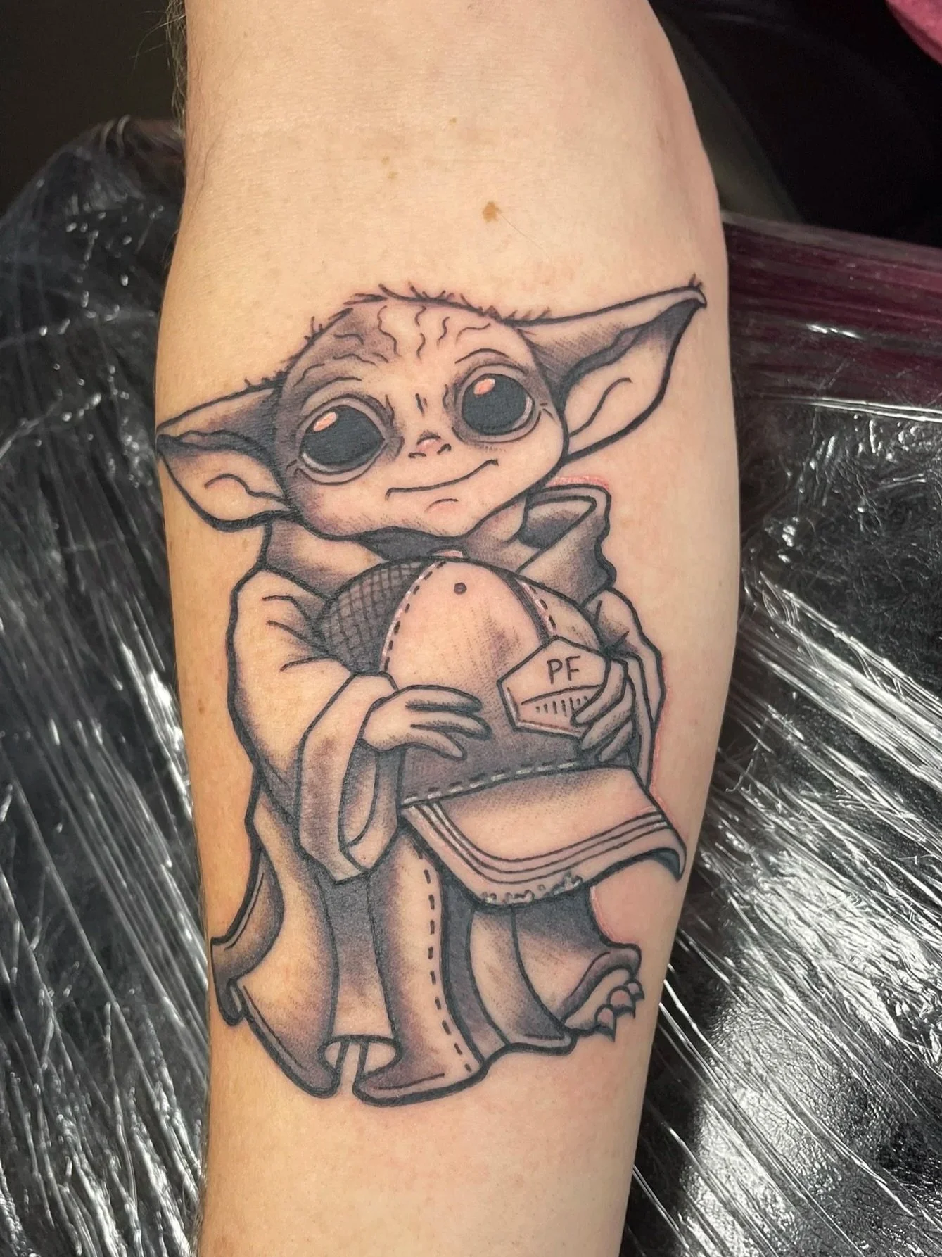 A tattoo of Baby Yoda holding a hat with memorial initials on it, in black ink and shading style, tattooed by Zing Phelps, in Cedar Rapids, Iowa