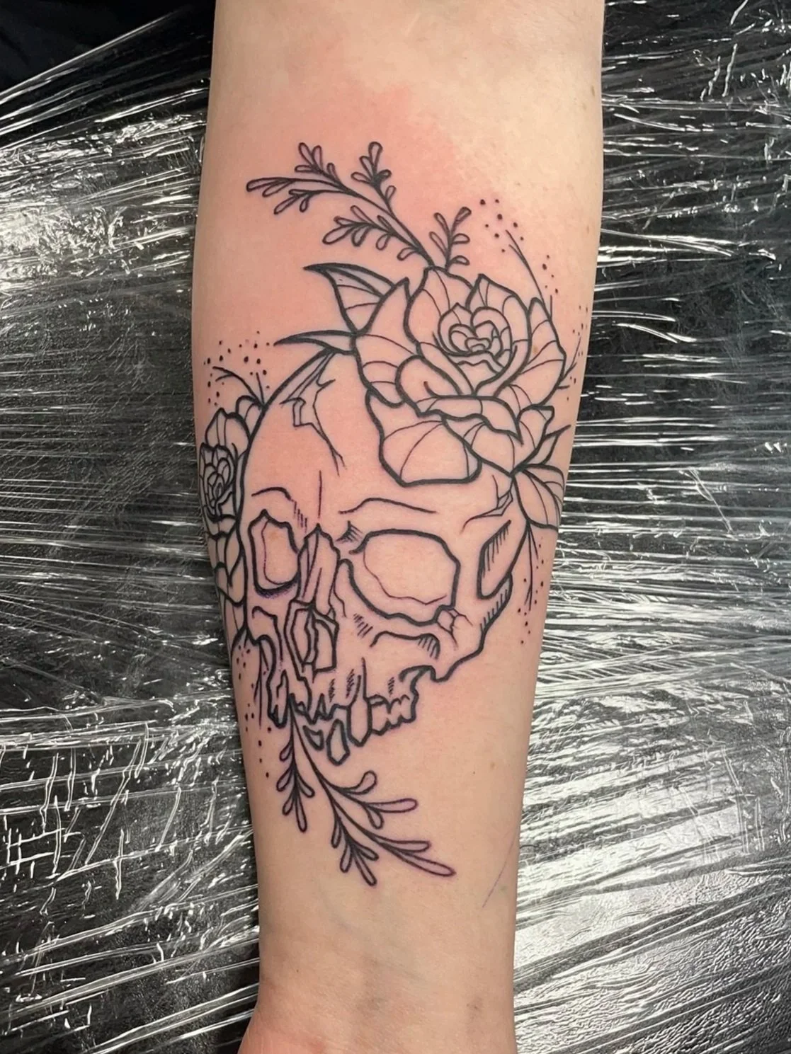 Black outline tattoo of a human skull with roses and leaves, with scattered small dots around the design on person's arm, tattooed by Zing Phelps, in Cedar Rapids, Iowa