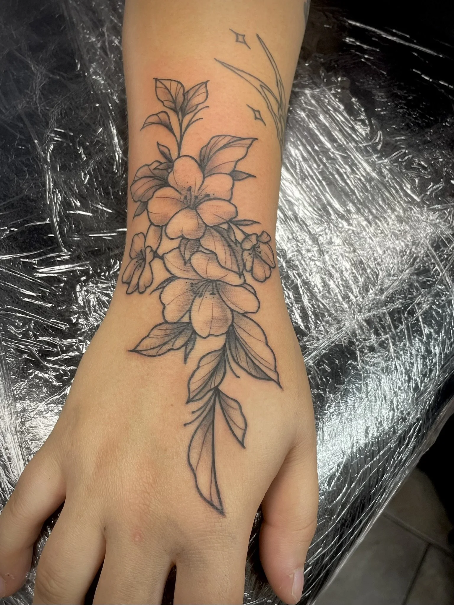 Black outline tattoo of blooming flowers and leaves on a person's hand, tattooed by Zing Phelps, in Cedar Rapids, Iowa