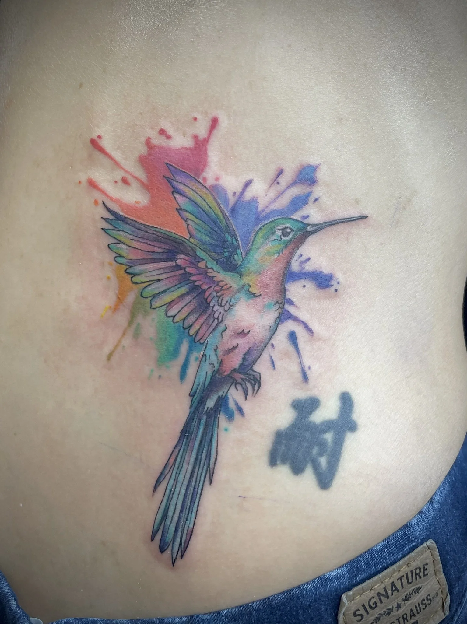 Colorful tattoo of a hummingbird with wings spread, surrounded by watercolor splashes in full rainbow colors, tattooed by Zing Phelps, in Cedar Rapids, Iowa