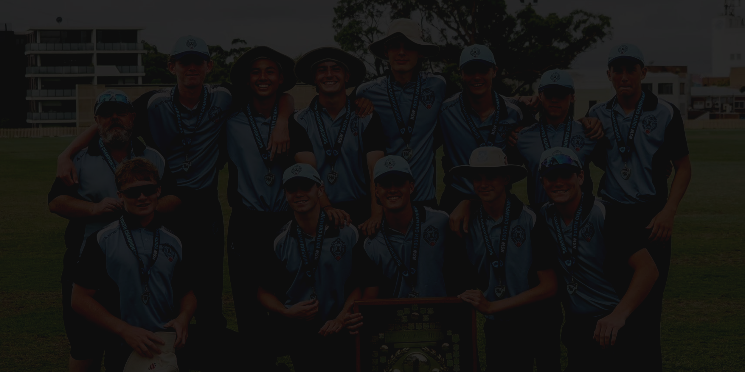 Cricket game being played on a grassy field on a clear day with players in white uniforms and tall light poles in the background.