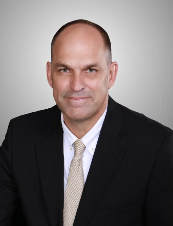A professional headshot of Dr. David Tomlinson wearing a black suit, white shirt, and beige tie, set against a neutral gray background.