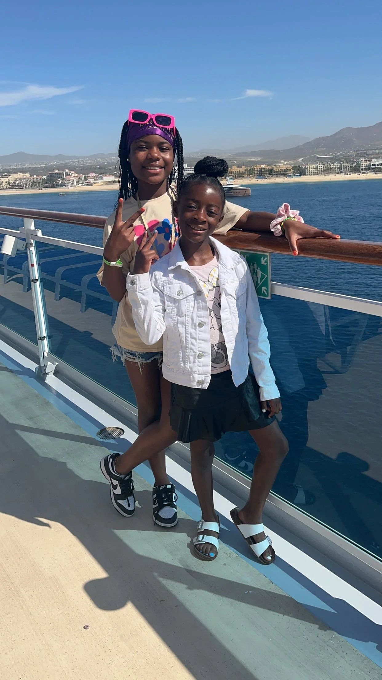 Two young girls posing together on a ship deck with a scenic water and city background. One girl is wearing a white jacket and black skirt, the other is in a tan t-shirt with colorful design and shorts. They are smiling and making peace signs.