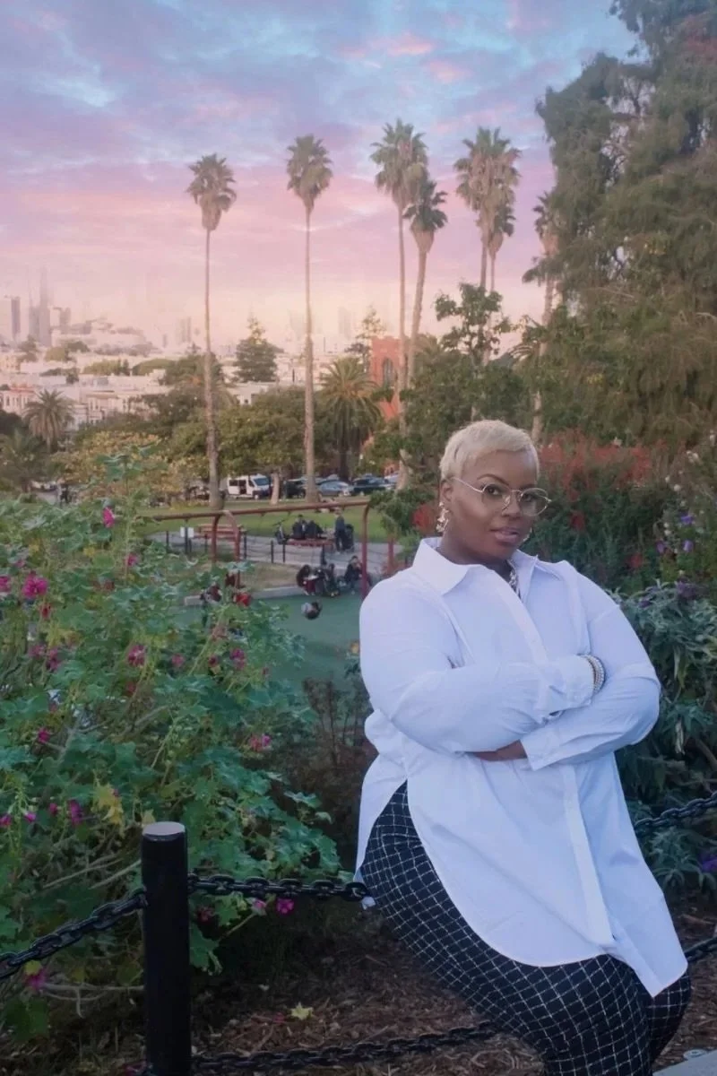 A woman with short blonde hair and glasses standing with arms crossed in a park. Background includes tall palm trees, a city skyline, and a colorful sunset sky.