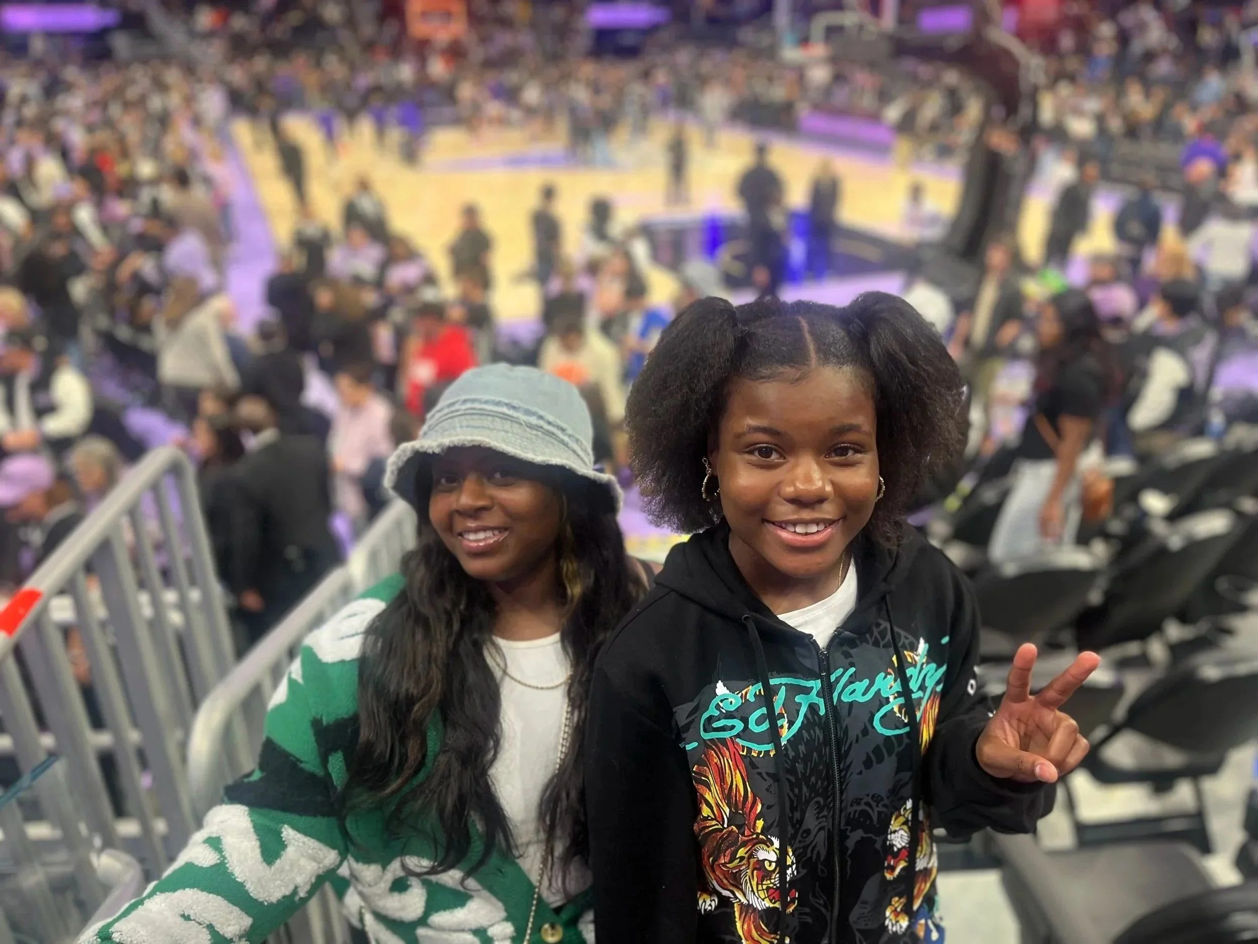 Two smiling young girls at a basketball game, one wearing a light blue bucket hat and green and white sweater, the other with puffy hair in a black hoodie, making a peace sign, in a crowded indoor arena.