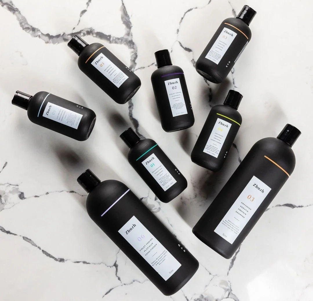 Collection of black bottles with white labels, arranged on a white marble surface, labeled as Zhusz hair care products including shampoo, conditioner, and scalp treatment.