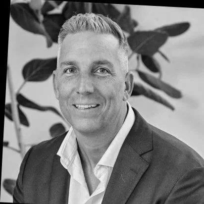 A black and white portrait of a man in a suit jacket and white shirt, smiling at the camera, with blurred leaves in the background.