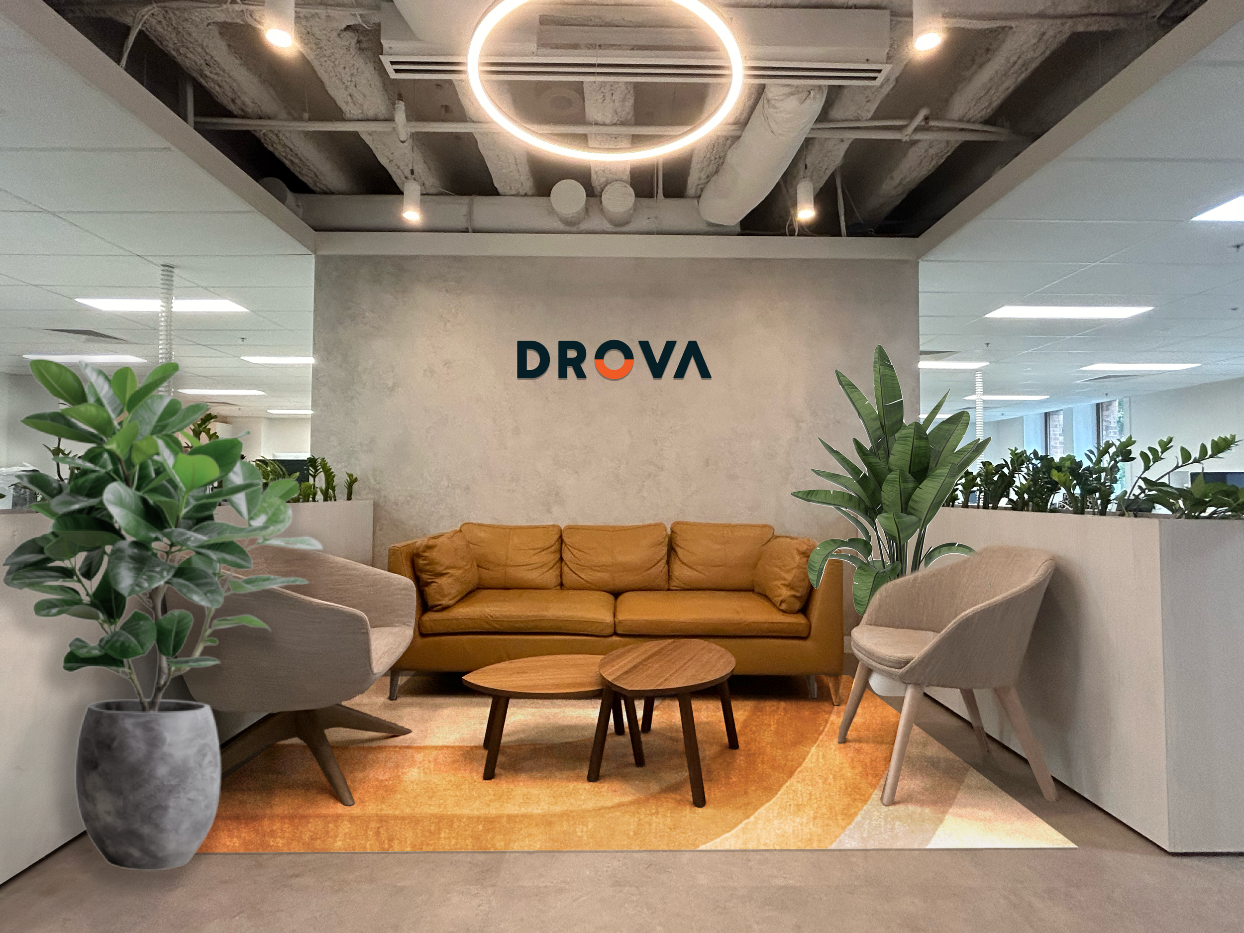 Branded Office Graphics - lounge area with a tan couch, two beige armchairs, and two round wooden coffee tables. Potted plants on either side of the seating area and a wall with the logo 'DROVA' in the background. Overhead circular light fixture.