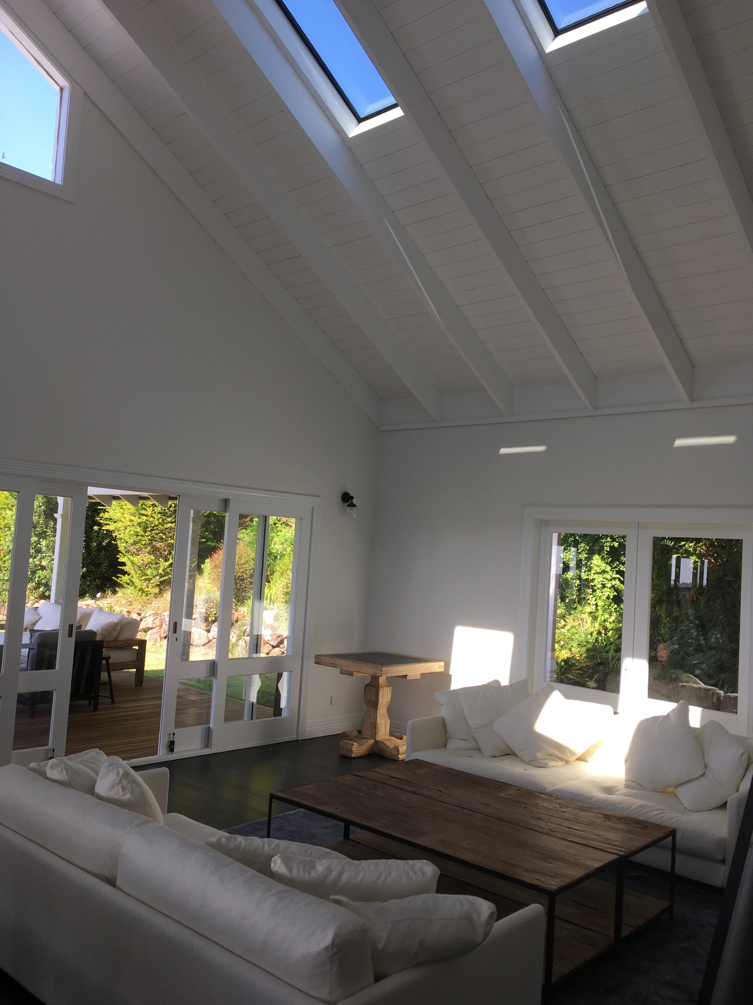 Bright living room with white sofas, a rustic wooden coffee table, and large windows and skylights allowing natural light to fill the space.