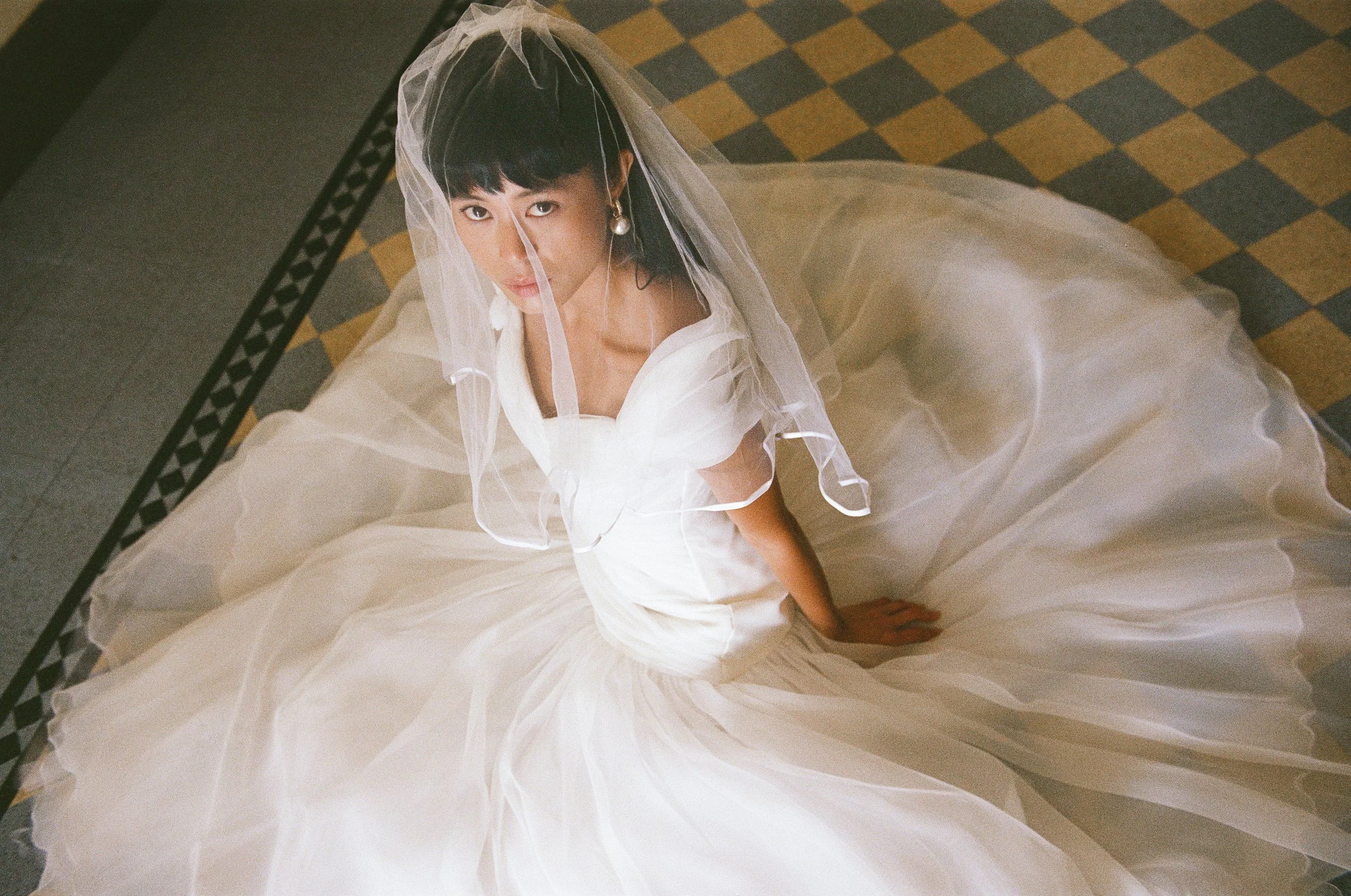 A woman in a white wedding dress with a veil, sitting on a checkered floor, looking up at the camera.