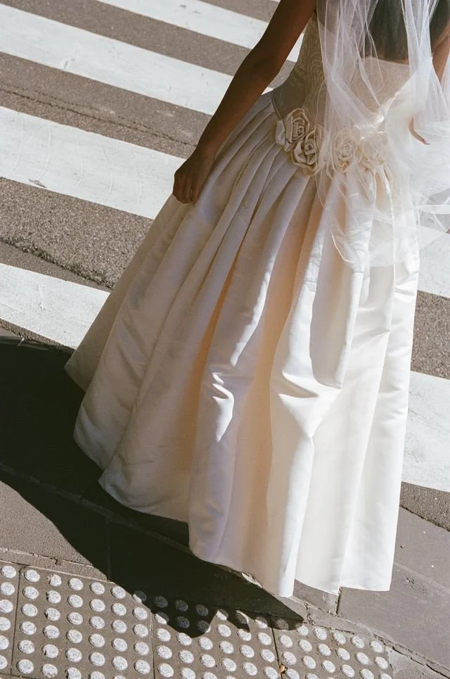 A woman wearing a white wedding dress with floral embellishments at the waist, standing on a street with pedestrian crossing lines.