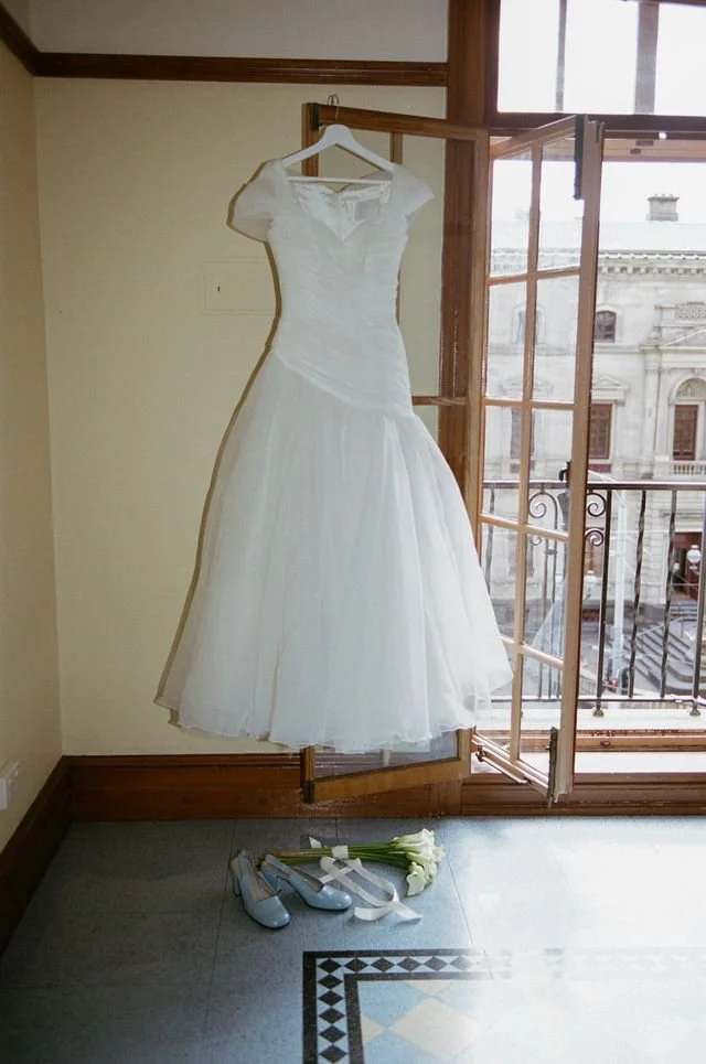 A white wedding dress hangs on a hanger inside a room near a large window with a balcony. On the floor below, there are a pair of light blue shoes, a bouquet of white flowers, and a ribbon.