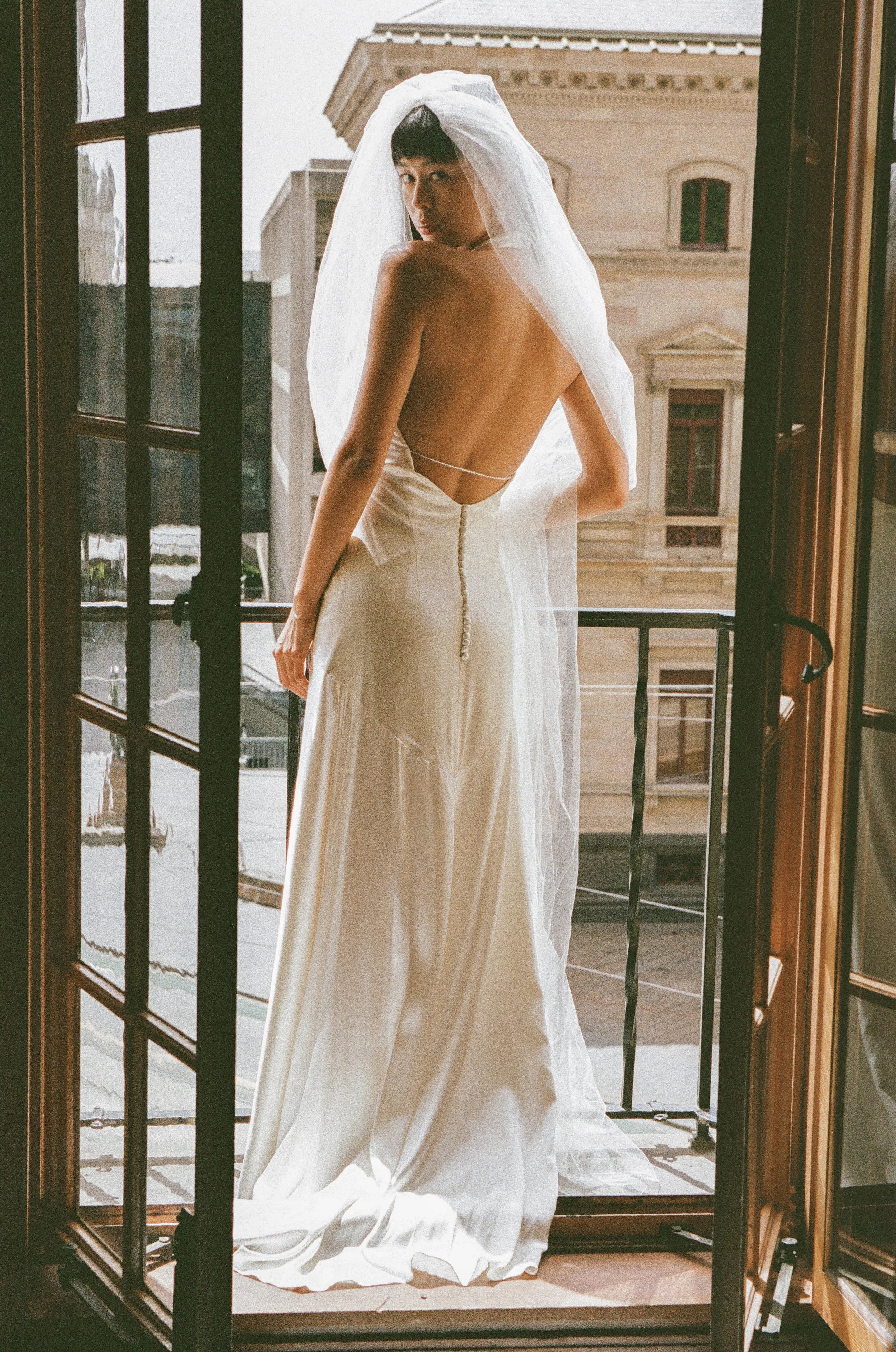 A woman in a strapless wedding dress with a long veil standing on a balcony, looking back over her shoulder. The city buildings are visible in the background.