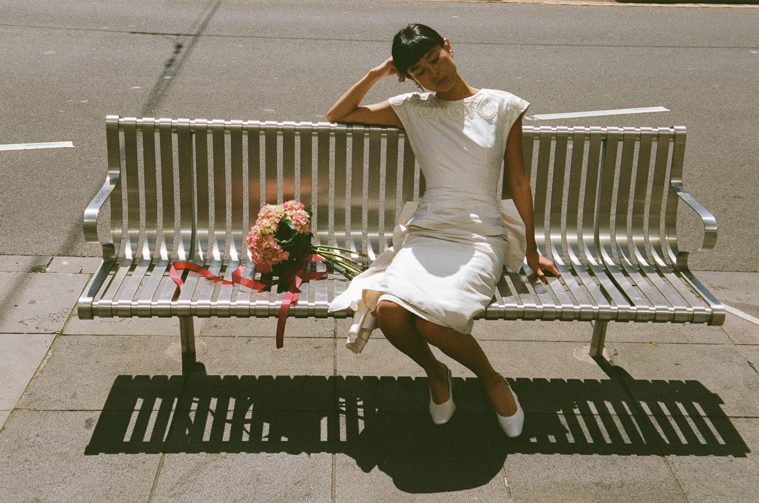 A woman in a white dress and white shoes sitting on a metal park bench with a bouquet of pink flowers beside her, on a sunny sidewalk.